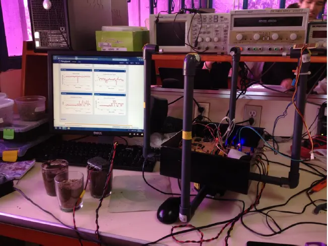 Lab bench with a weather-station prototype, soil sensor samples, and a monitor showing live telemetry charts